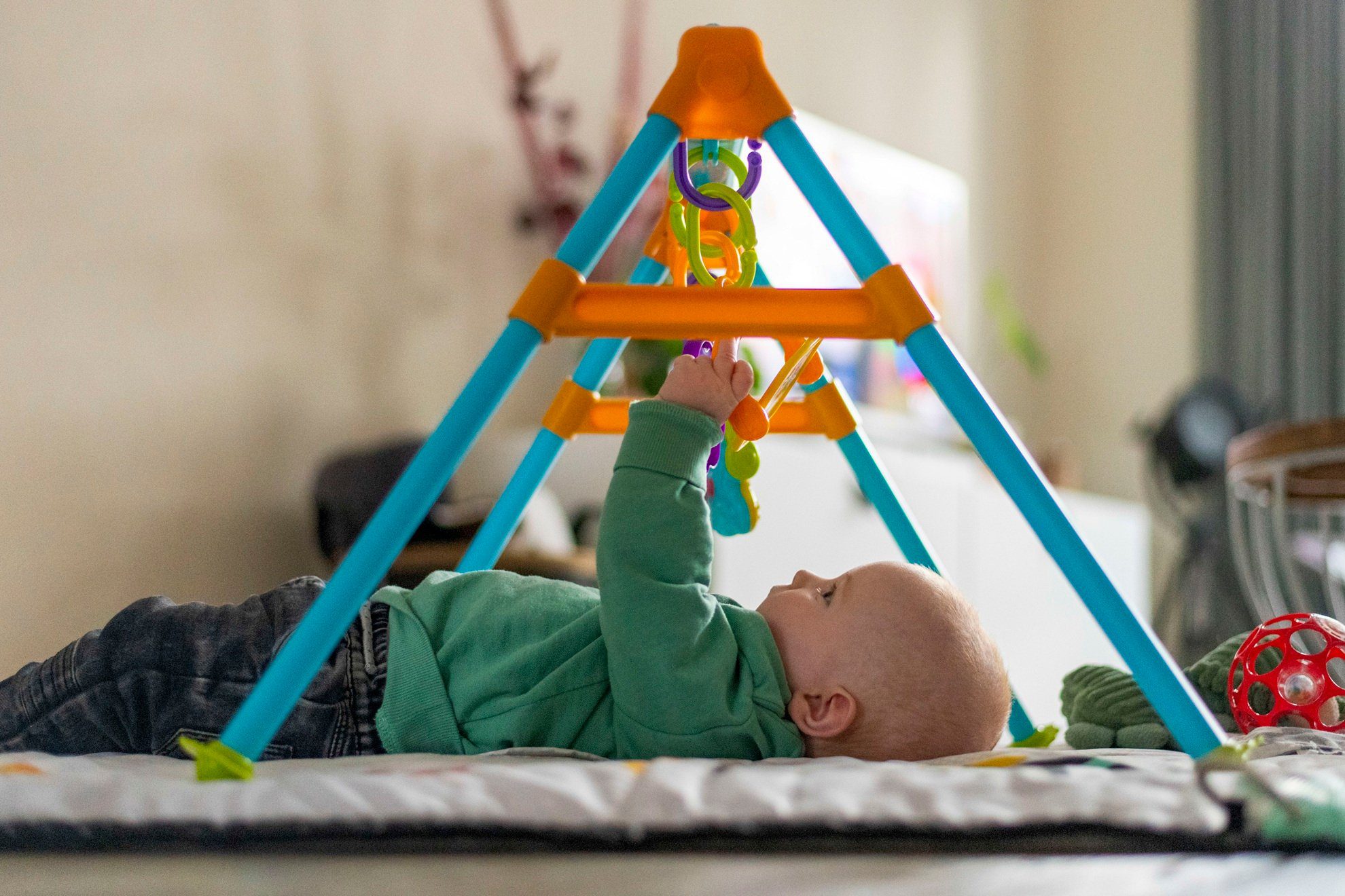 A baby lies on a mat, reaching for colorful toys on an orange and blue baby play gym.