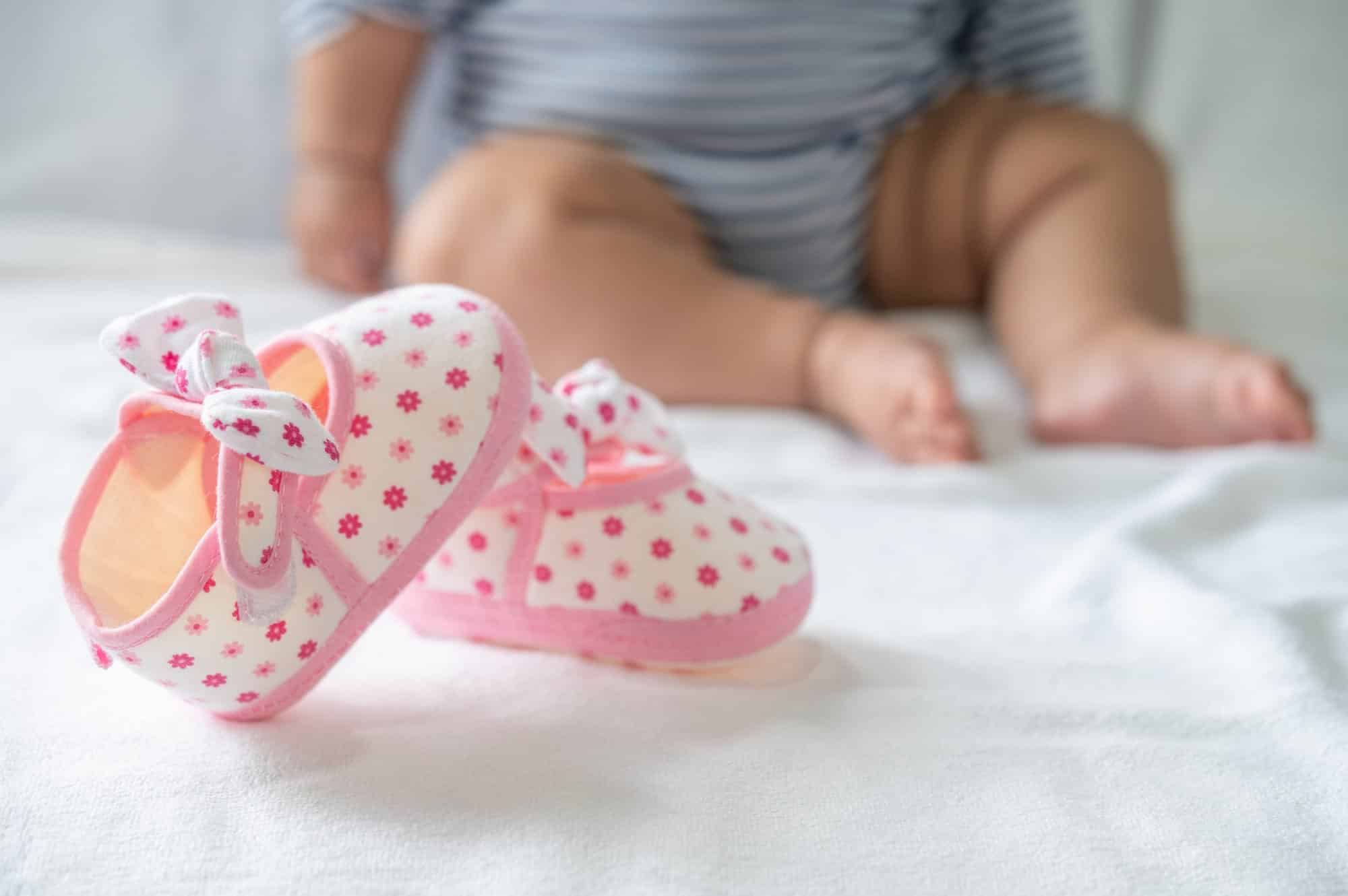Close-up of pink infant girl shoes with a floral pattern and bows, a blurry baby in background.