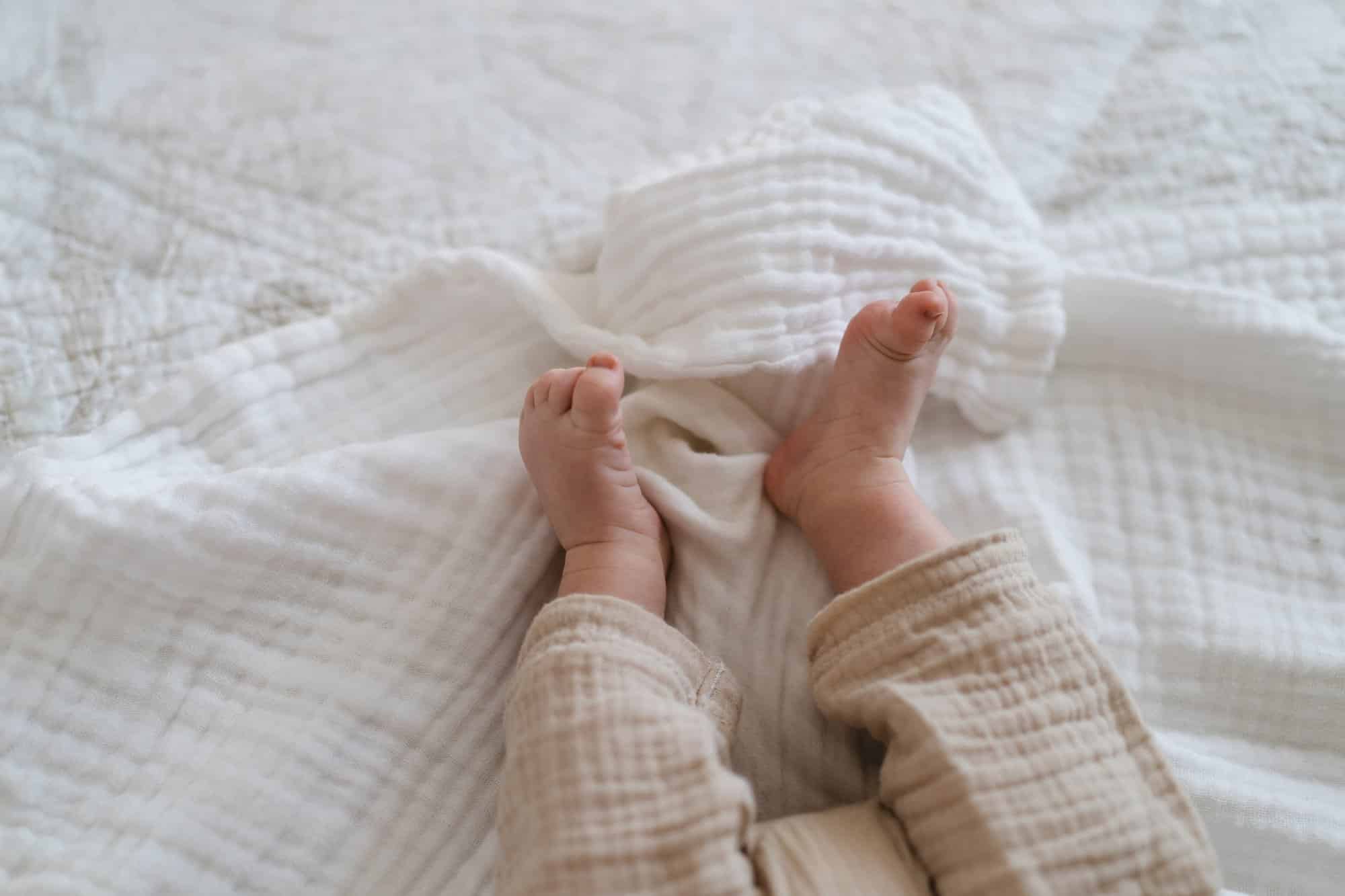 Close-up of a baby's bare feet resting on a soft baby muslin blanket, wearing light beige pants.